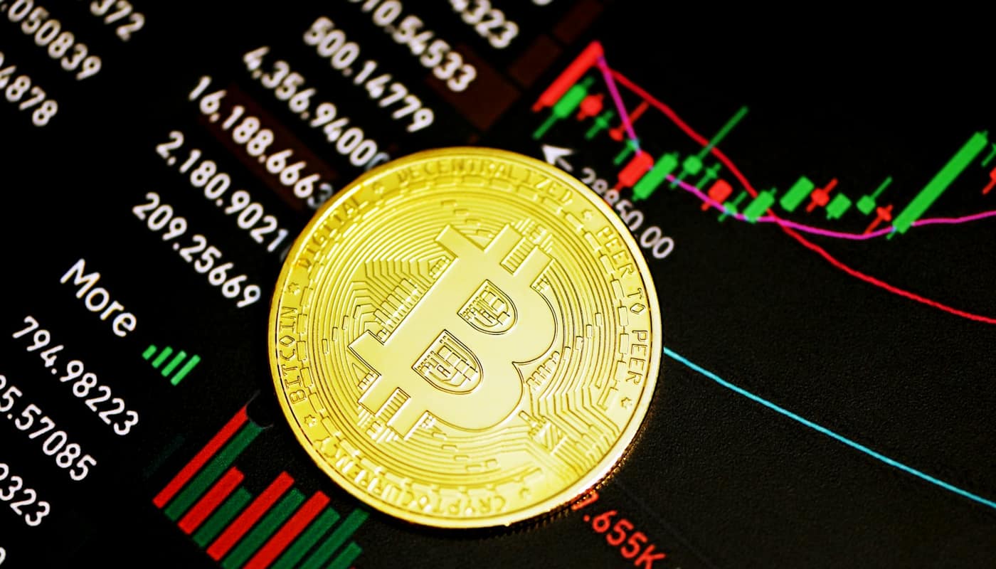 A gold bitcoin coin over a screen of numbers and a price chart