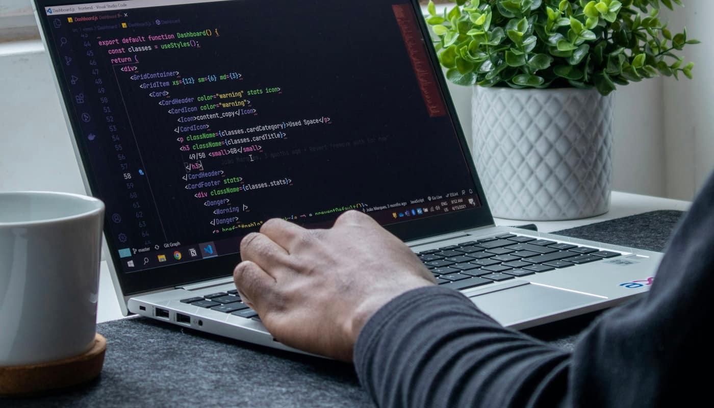 A person coding on a laptop beside a plant and a cup